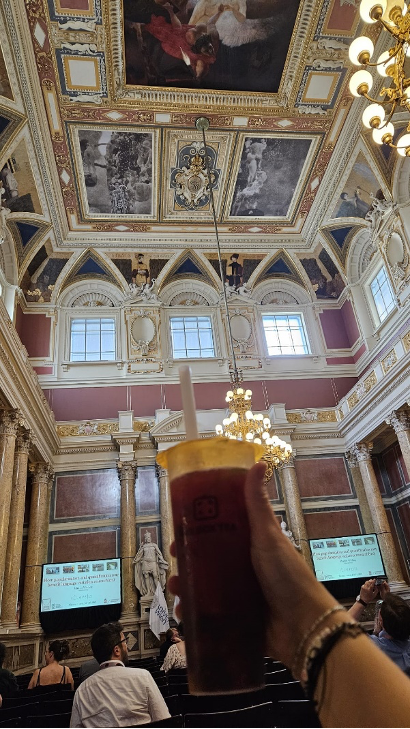 A hand with bracelets is holding a plastic cup with a straw containing a berry-colored liquid with boba. In the background, you can see an old building with high ceilings, pompous pillars, a chandelier, a statue, and two screens that display the same title slide of the plenary’s slideshow. A few of the chairs are already occupied.